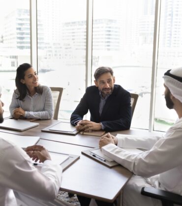 A group of people, dressed in professional attire, sitting around a conference table in a modern office setting with a cityscape visible through the windows.