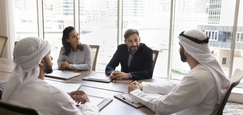 A group of people, dressed in professional attire, sitting around a conference table in a modern office setting with a cityscape visible through the windows.