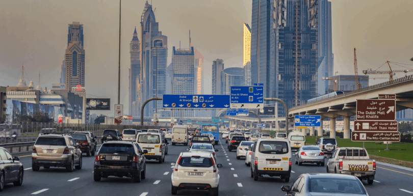 Heavily congested urban highway in Dubai, with numerous vehicles of different sizes and types stuck in traffic, surrounded by a backdrop of tall skyscrapers and modern high-rise buildings