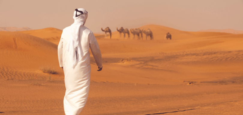 Emirati man in traditional dress walking in the desert towards a herd of camels, symbolizing enforcement and seizure of camels in the UAE